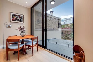 Un comedor con una mesa y sillas de madera, un florero con flores y un ventanal con vistas a un edificio y vegetación.