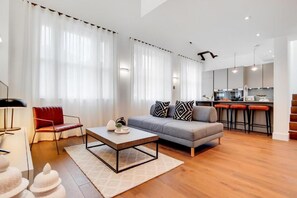 A modern living room with a sofa, a coffee table, and a red chair. There is a kitchen area with a bar stool and a staircase. - Spacious Apartments in Borough London Bridge by Short Stories London (London)