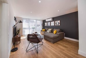 A modern living room with a sofa, a coffee table, and a chair. There is a window with blinds, a lamp, and artwork on the wall. - Spacious Apartments in Borough London Bridge by Short Stories London (London)