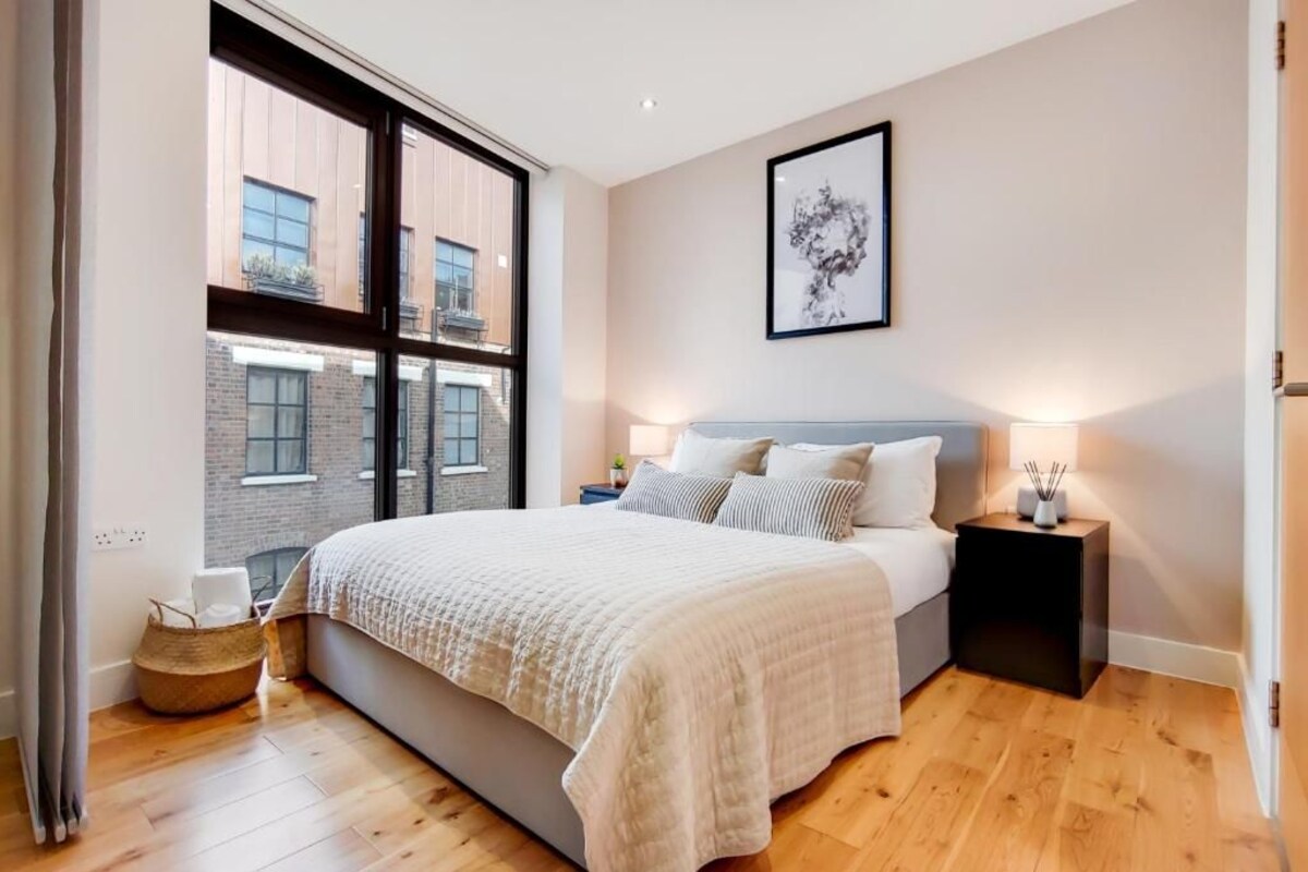 A bedroom with a large bed, a window with a view of buildings, a framed artwork on the wall, and a bedside table with a lamp.
