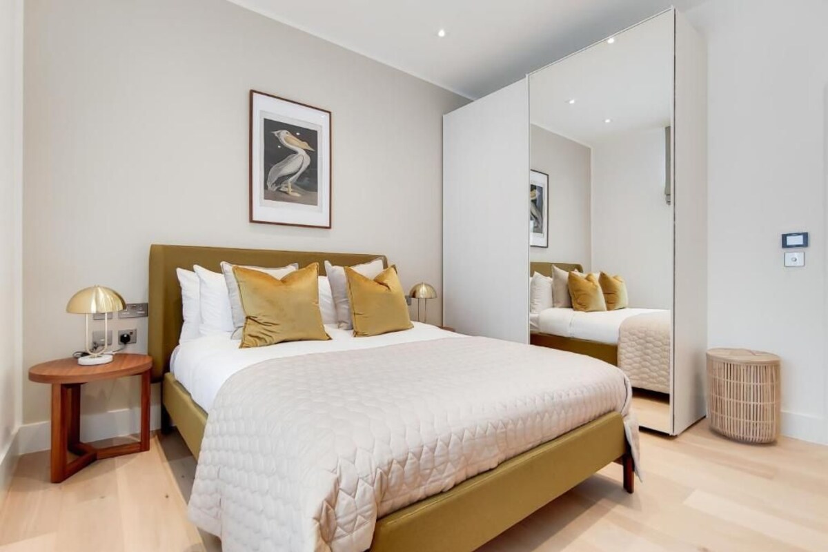A modern bedroom with a large bed, bedside lamps, a framed artwork on the wall, and a mirror reflecting the bed.