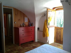Una habitación con una cama, un aparador rojo, un ventanal con cortina amarilla y una puerta de madera.