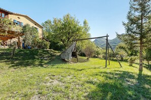 A rural setting with a wooden swing set, a stone structure, and a building with blue shutters.
