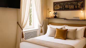 A hotel room with a bed, curtains, a painting, a shelf with books, and a television mounted on the wall.