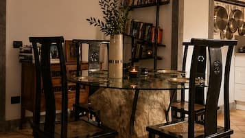 A dining area with a glass table surrounded by wooden chairs, a potted plant, and a bookshelf with books and decorative items.