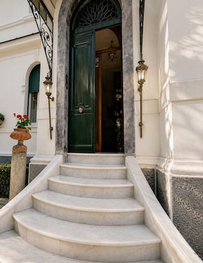 A set of marble stairs leading to a green door with a wrought iron railing and two wall-mounted lamps. - Casa Rubinacci (Napoli)