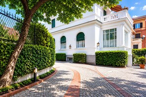 A white building with a cobblestone pathway leading to the entrance, surrounded by greenery and a fence.