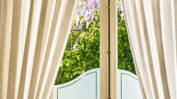 A view through a window with curtains, a white door with a brass handle, and a glimpse of purple flowers and greenery outside.