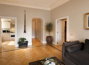Amplia sala de estar con piso de madera en espiga, un sofá gris oscuro y una mesa de centro con flores. Se ve una cocina abierta con gabinetes de madera y un microondas al fondo.