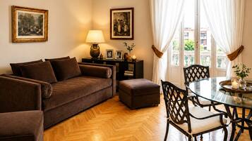 Una sala de estar con un sofá marrón, una mesa de vidrio y sillas, una lámpara, cuadros enmarcados y un ventanal con cortinas.