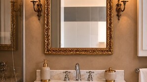 A bathroom with a large ornate mirror, two wall-mounted sconces, a white marble sink with gold faucets, and a towel rack. - Casa Rubinacci (Napoli)