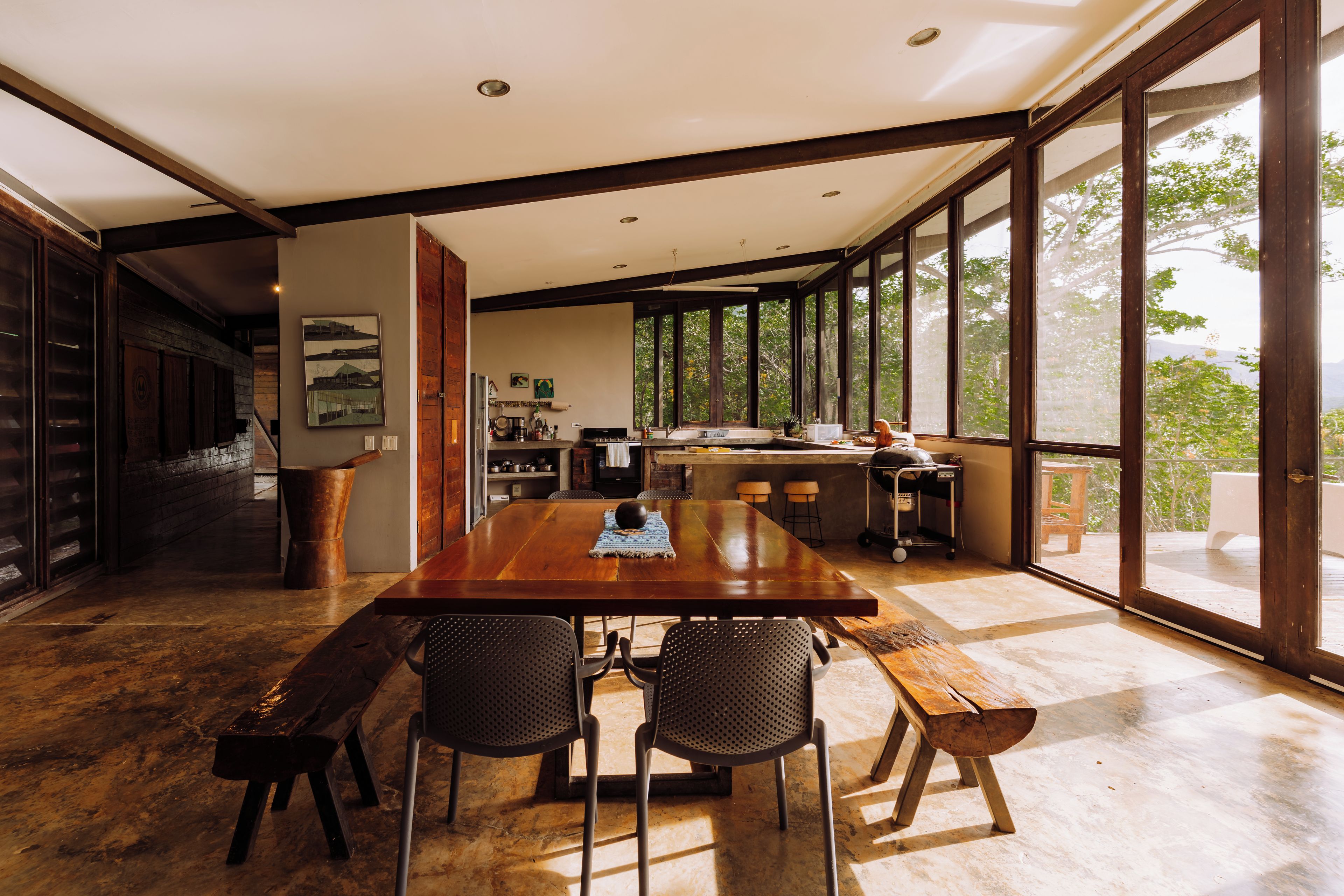 A spacious dining area with a large wooden table and chairs, a kitchen in the background, and large windows offering a view of the outdoors.
