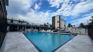 A swimming pool with a tiled deck, lounge chairs, and a building in the background.