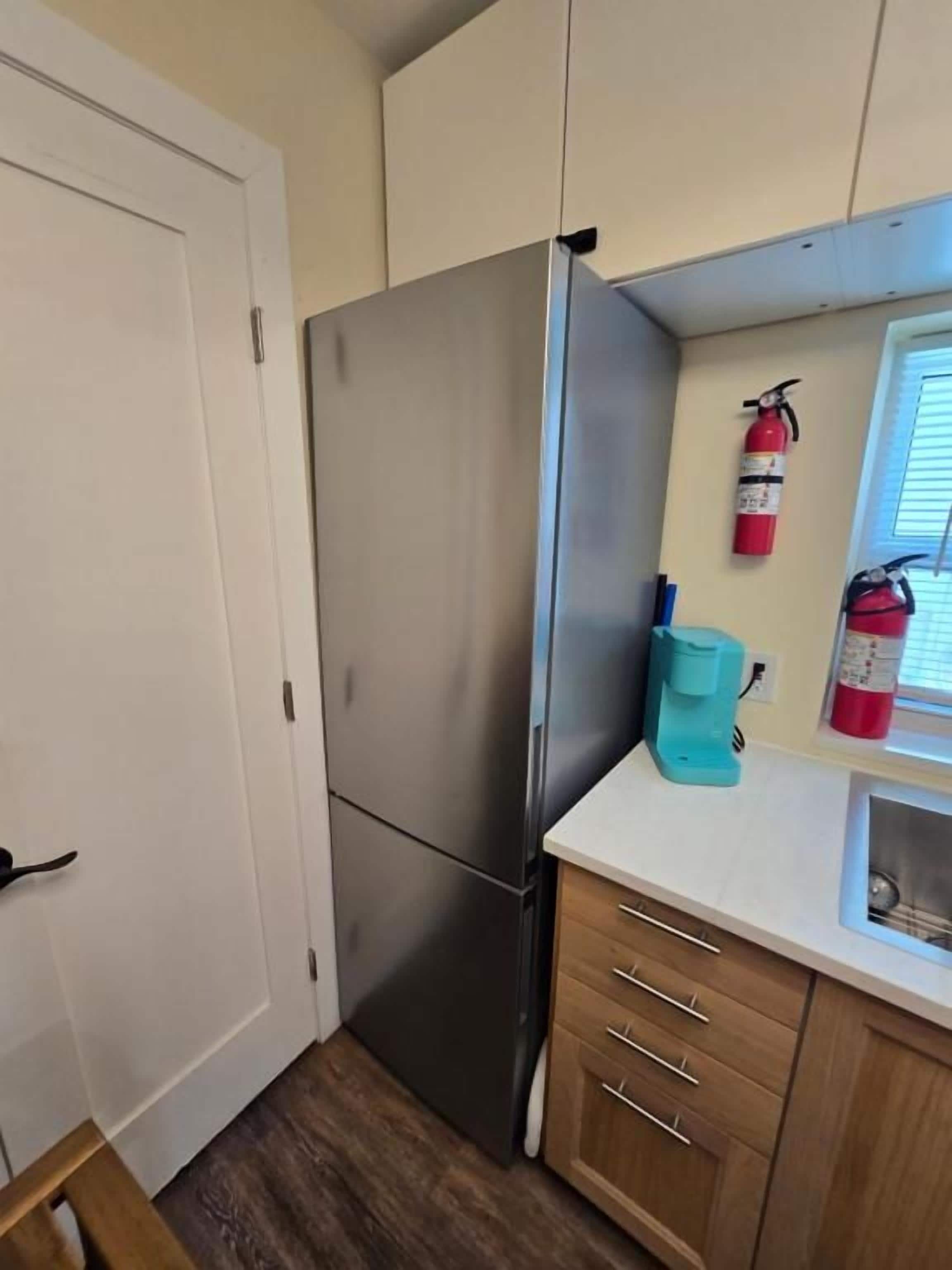 Una cocina compacta con refrigerador de acero inoxidable, armarios de madera, una encimera, extintor de incendios y ventana.