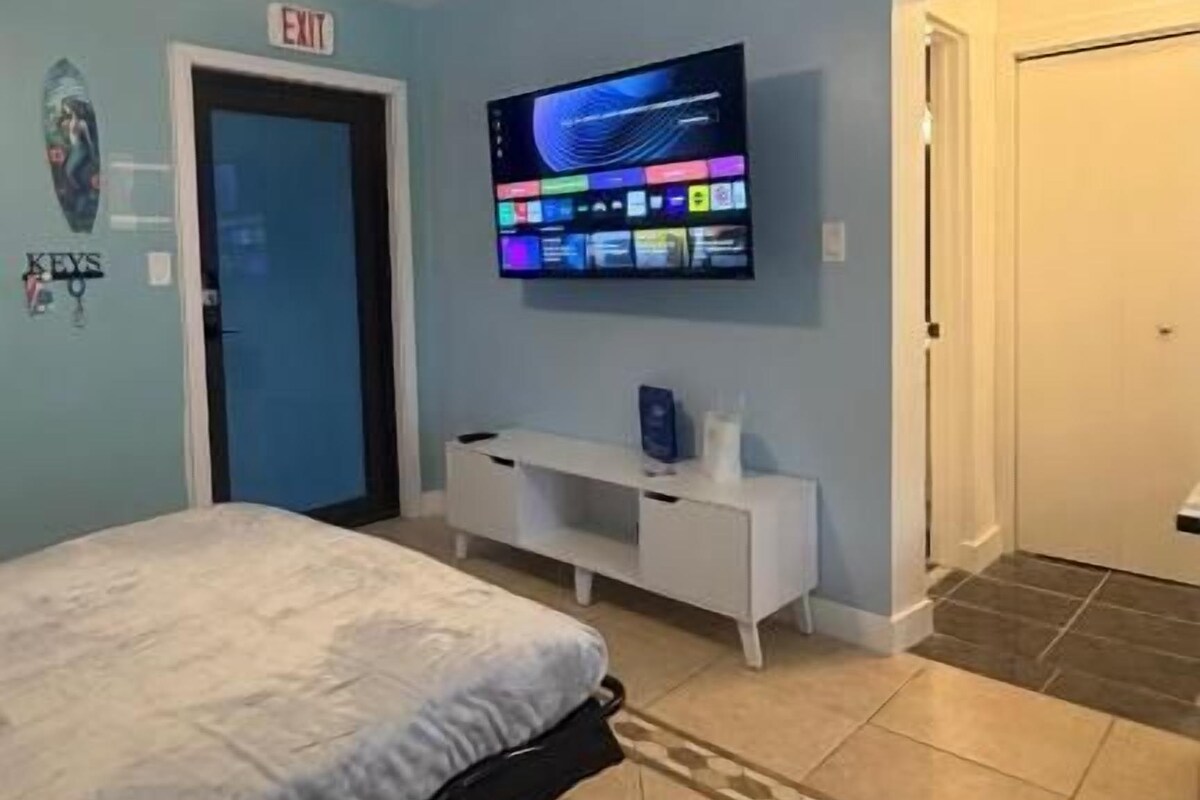 Ein Schlafzimmer mit einem Bett, einem an der Wand montierten Fernseher, einem weißen Schminktisch und einer Tür mit einem Ausgangsschild.