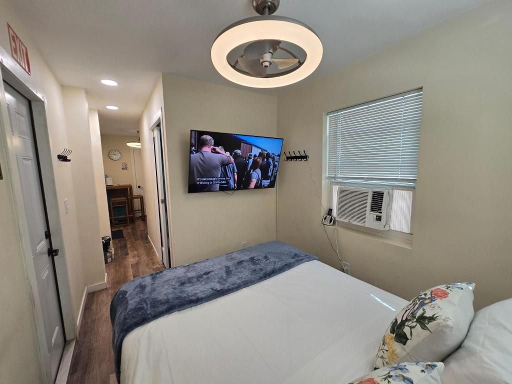 Habitación de hotel con cama, televisor montado en la pared, ventilador de techo y ventana con persianas.