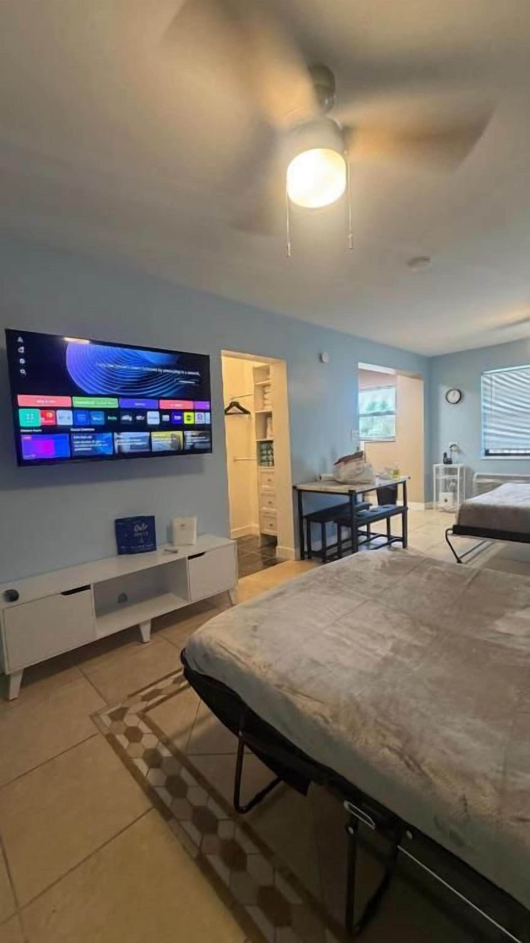 Un dormitorio con cama, televisión montada en la pared, escritorio con silla y ventana con persianas.