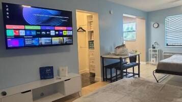 Un dormitorio con cama, televisión montada en la pared, escritorio con silla y ventana con persianas.
