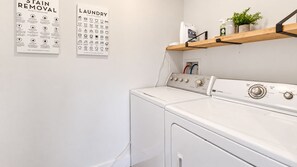 A laundry area with a washing machine and dryer, a wall-mounted shelf with cleaning supplies, and two instructional posters on stain removal and laundry procedures. - 3BR Modern Retreat | Hot Tub | Near 4 Ski Resorts (South Salt Lake)
