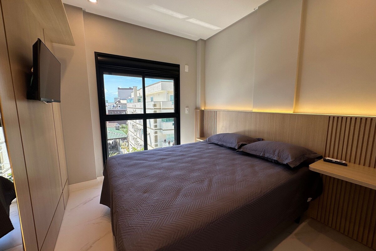 A modern bedroom with a bed, a TV mounted on the wall, and a window with a city view.