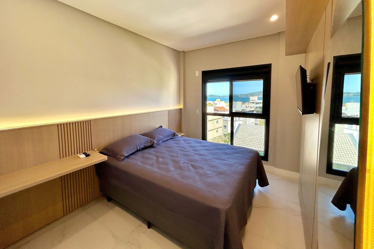 A modern bedroom with a bed, bedside table, and a large window offering a city view.