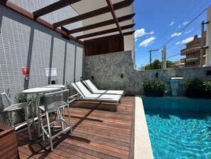 A poolside area with lounge chairs and a table, a stone wall, and a tiled building facade.
