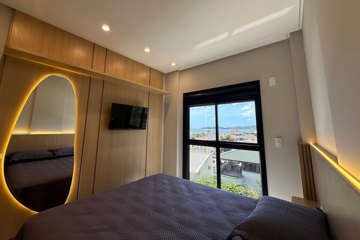 A modern bedroom with a bed, a TV mounted on the wall, and a window with a view of buildings and a clear sky.