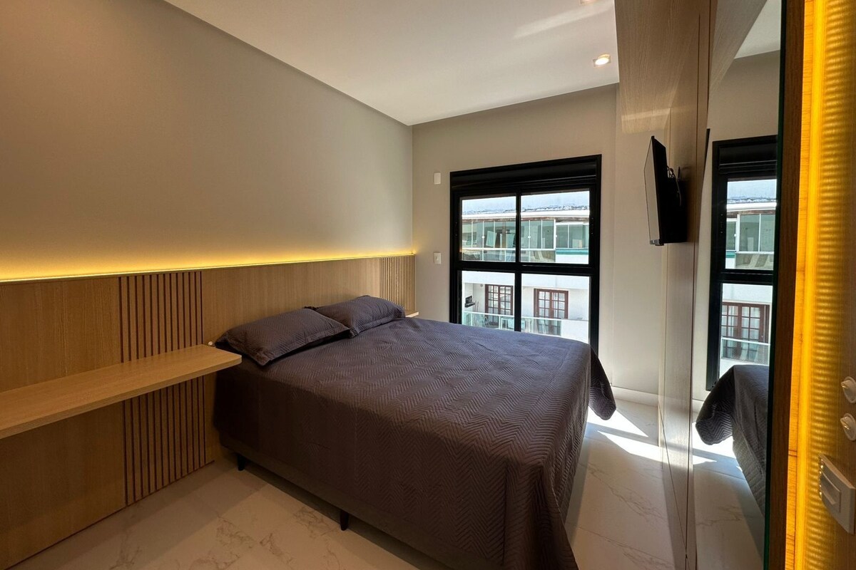 A modern bedroom with a bed, a TV mounted on the wall, and a window with a view of buildings.