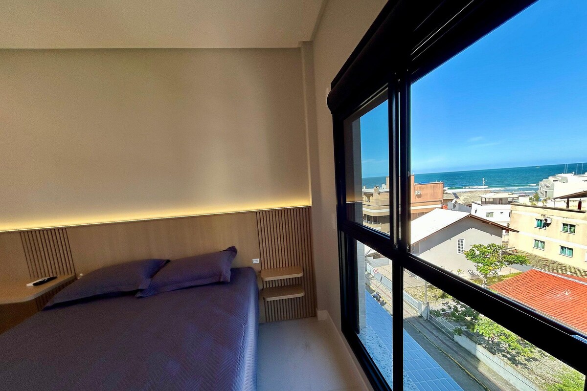 A bedroom with a large window, a bed, and a view of the sea and buildings.