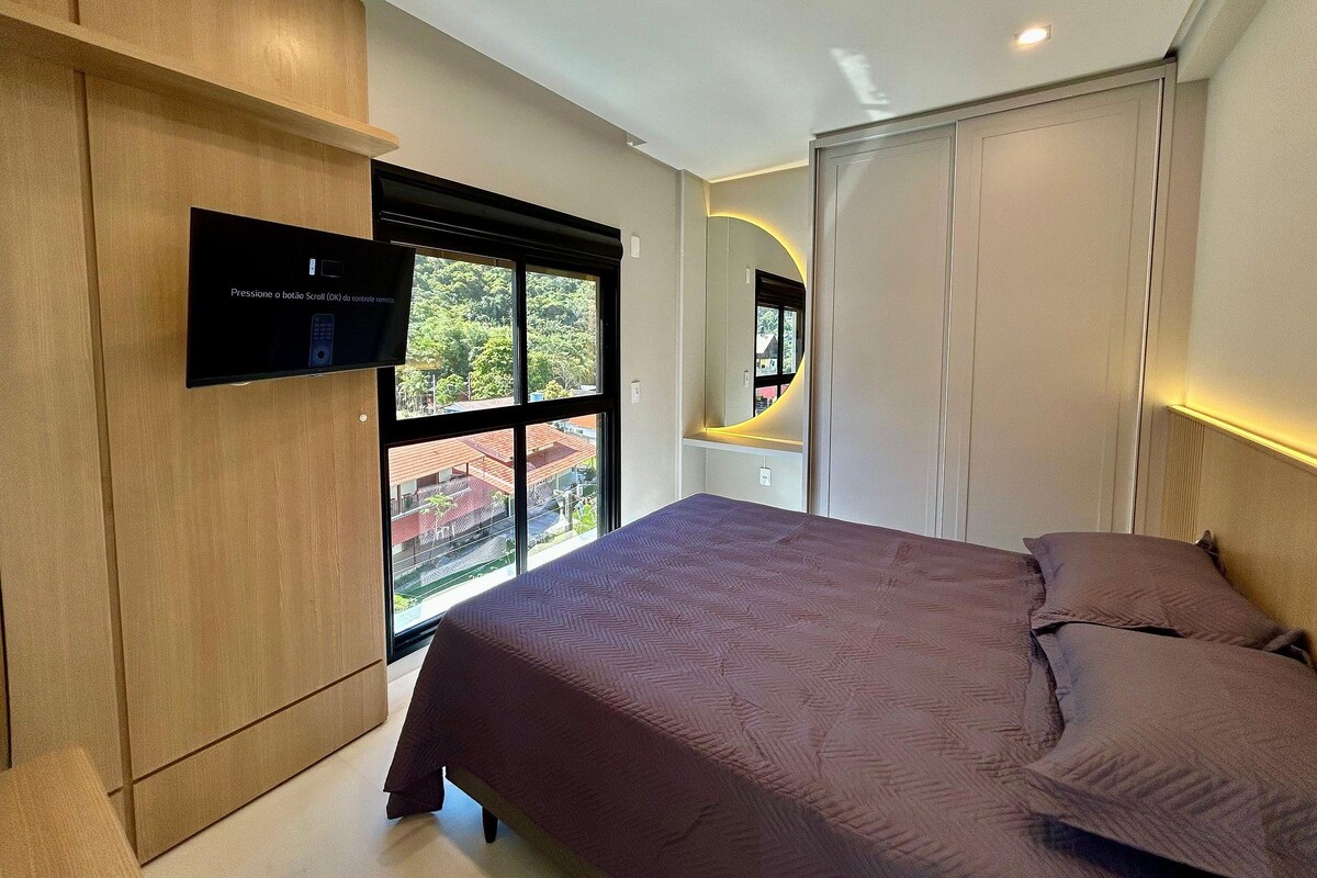 A modern bedroom with a bed, a TV mounted on the wall, and a large window offering a view of the outside.