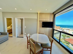 A modern living room with a dining area, a TV mounted on the wall, and a view of the ocean. - Mirante do Oceano Hospedaria (Bombinhas)