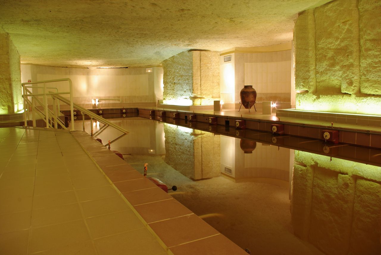Una piscina interior con suelo de baldosas, una fuente central y una pared de piedra en un lateral.