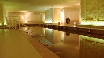 Una piscina interior con suelo de baldosas, una fuente central y una pared de piedra en un lateral.