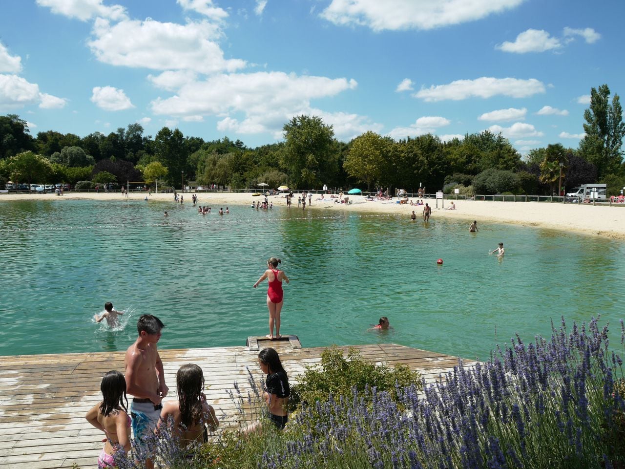 Een zwemgedeelte met een houten terras, mensen in het water en een strand op de achtergrond.