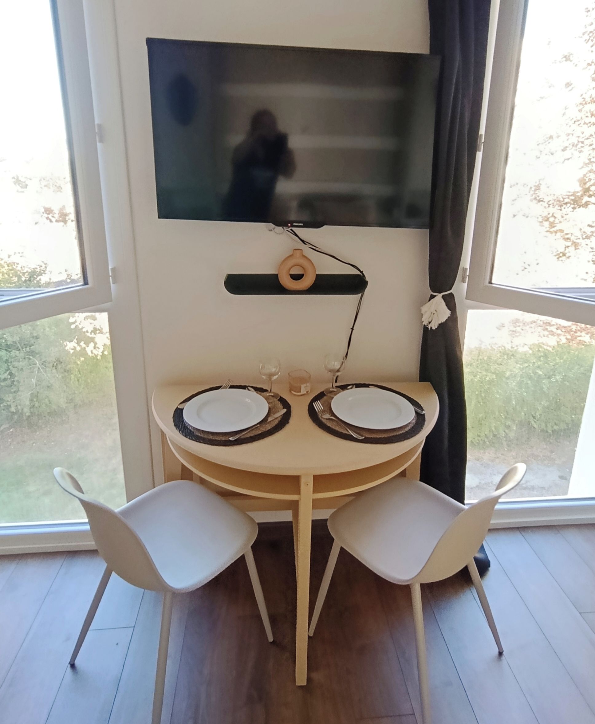 Un comedor con una mesa redonda, dos sillas blancas, un televisor montado en la pared y vistas al exterior a través de amplias ventanas.