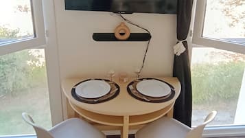 Un comedor con una mesa redonda, dos sillas blancas, un televisor montado en la pared y vistas al exterior a través de amplias ventanas.