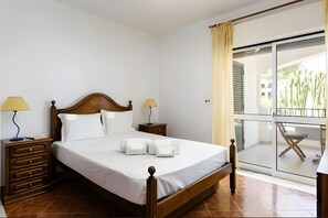 A hotel room with a wooden bed, two bedside lamps, a nightstand, and a view of a balcony with a chair. - Apartment 'Vilamoura Garden View' with Shared Pool, Wi-Fi and Air Conditioning (Quarteira)