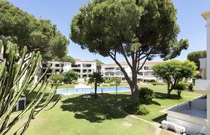 A residential area with a swimming pool, multiple-story buildings, and a large tree in the foreground. - Apartment 'Vilamoura Garden View' with Shared Pool, Wi-Fi and Air Conditioning (Quarteira)