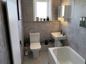 A modern bathroom with a toilet, sink, and bathtub. There are potted plants on the windowsill and a window above the toilet. - Sea View Luxury Apartment Sleeps 5 - Fast Wifi - off Street Parking - Contractors Dream (Sheerness)