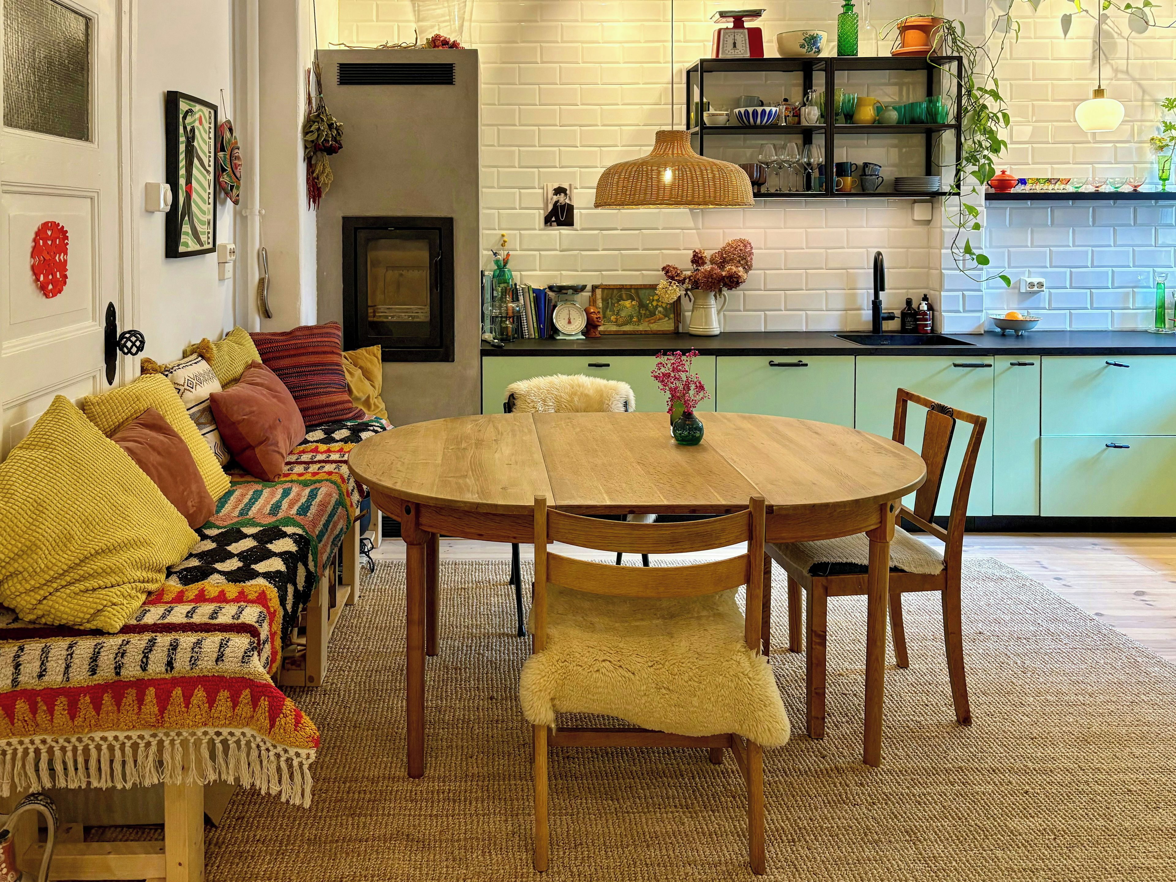 Een gezellige woonkamer met een open haard, een eettafel en een keukenhoek met een groen kastje en een hanglamp.