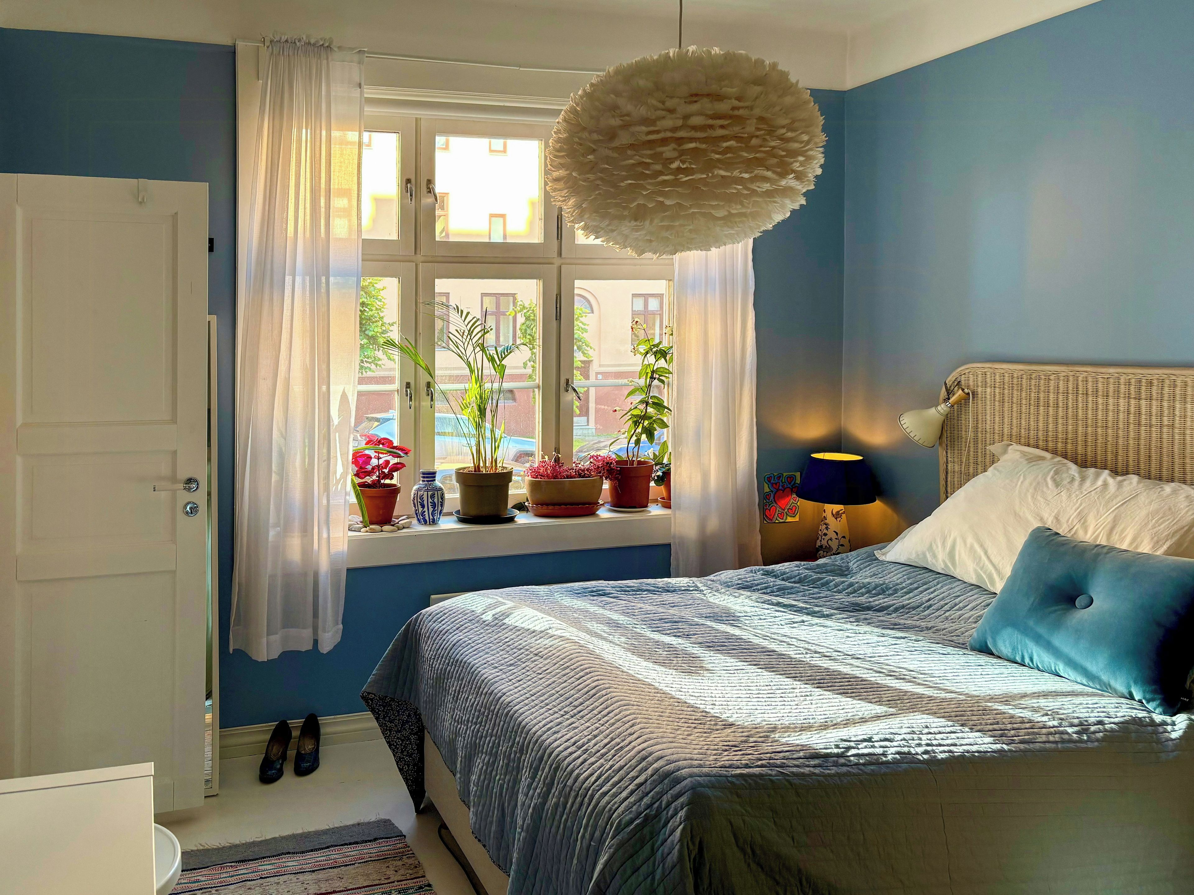 Un dormitorio con cama, ventana con cortinas, lámpara colgante y plantas en el alféizar.