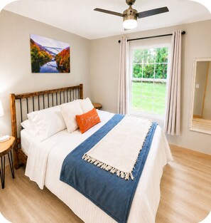 Quarto com cama, ventilador de teto, janela com cortinas, mesa de cabeceira e quadro na parede.