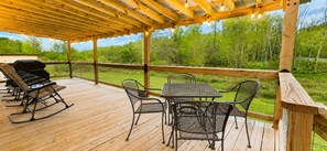 Um deck externo com pérgola de madeira, cadeiras de metal, uma mesa e uma churrasqueira.