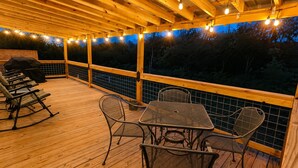 Een terras buiten met een houten vloer, metalen stoelen, een tafel en lichtslingers.