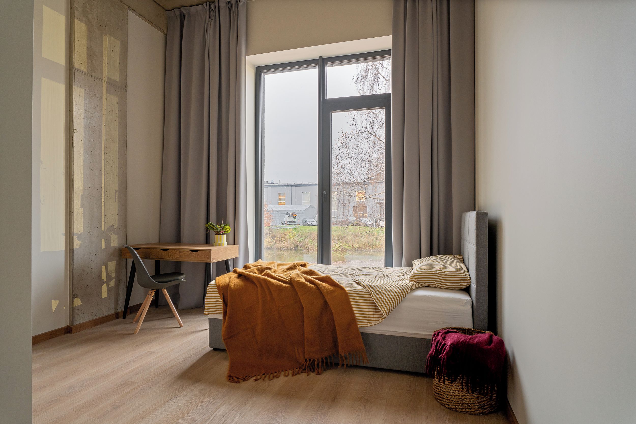 A bedroom with a bed, a desk, and a window with a view of a building and trees.