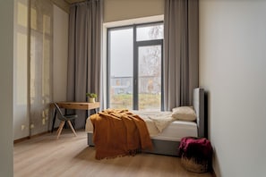 A bedroom with a bed, a desk, and a window with a view of a building and trees. - Uniflow Apartments by Rixwell (Rīga)