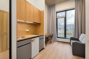 A compact kitchen with wooden cabinets, a built-in dishwasher, and a window with curtains. - Uniflow Apartments by Rixwell (Rīga)