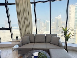 A modern living room with a large window, a grey sofa, a coffee table, and a potted plant.