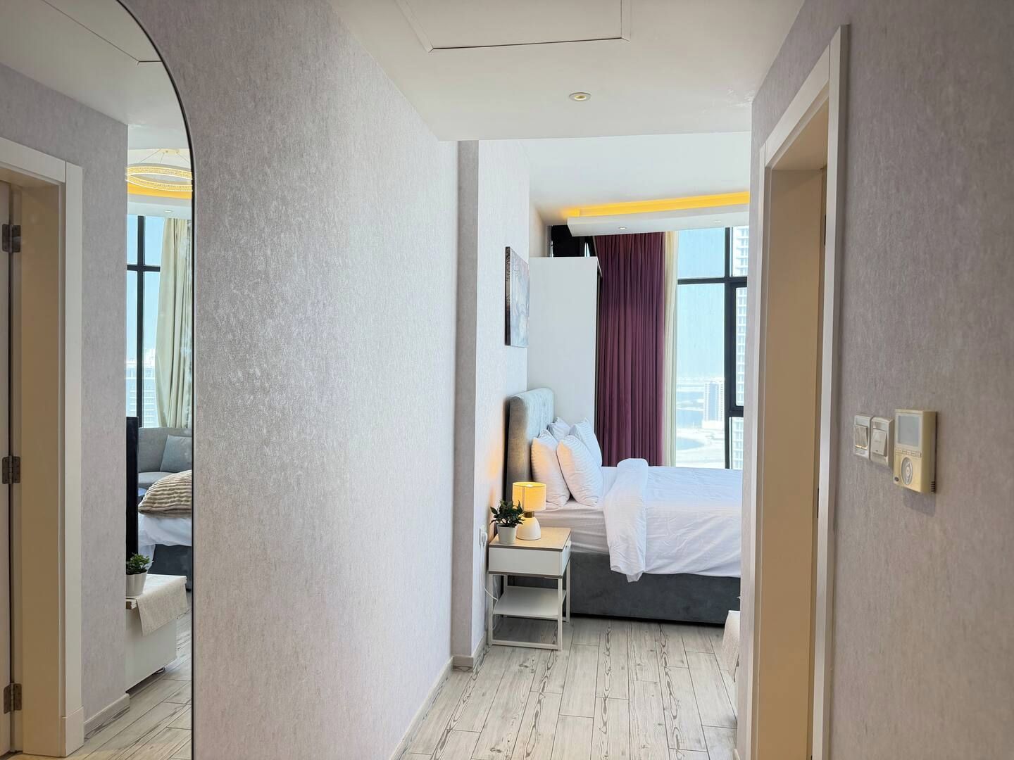 A hotel room with a bed, bedside table, lamp, and a view of the cityscape through large windows.