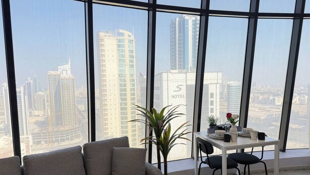 A modern hotel room with a large window offering a city view, a sofa, a small table with a vase of flowers, and a potted plant. - High-Floor Sea View Studio - Seef Area F296O (Seef)